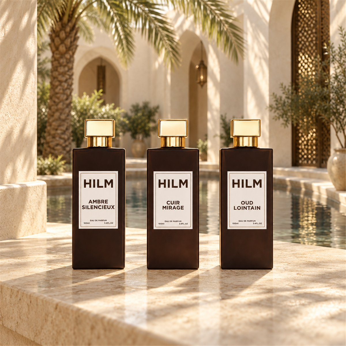 Three HILM perfume bottles Ambre Silencieux, Cuir Mirage and Oud Lointain, in dark amber glass with gold caps are lined up on a stone patio in front of a resort-style pool and archway.