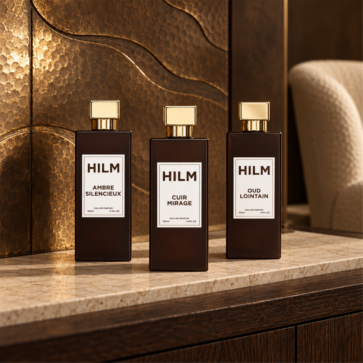Three dark amber glass perfume bottles of Ambre Silencieux, Cuir Mirage and Oud Lointain, with gold caps and white labels reading HILM, arranged on a marble countertop against a textured metallic bronze wall.