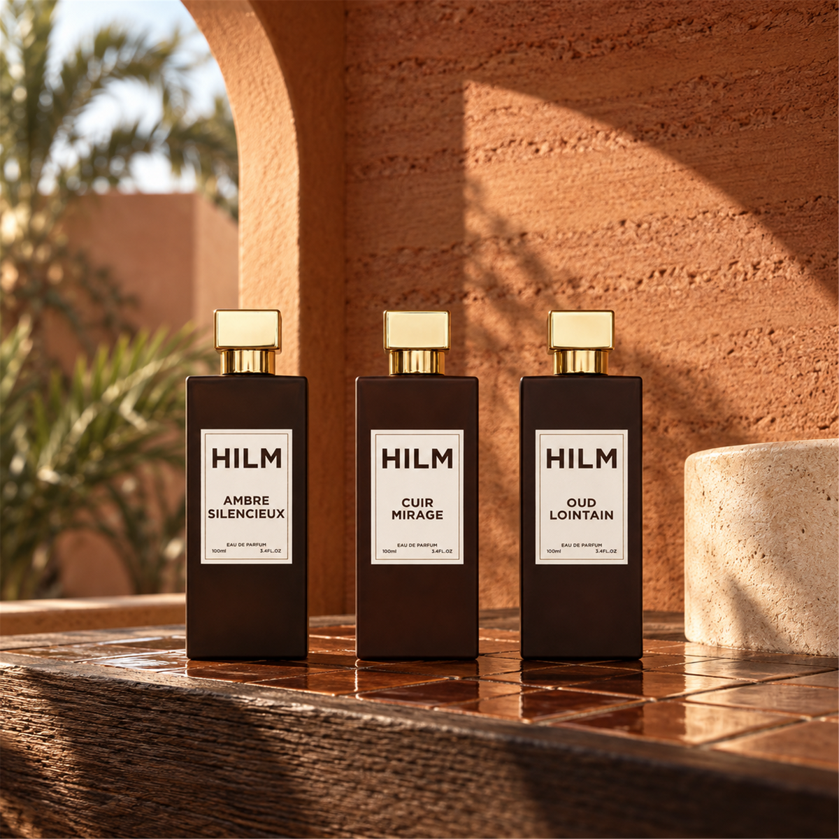 Three HILM perfume bottles, Ambre Silencieux, Cuir Mirage, and Oud Lointain, in dark amber glass with gold caps and white labels, arranged on a wooden surface against a warm terracotta wall with outdoor shadows.